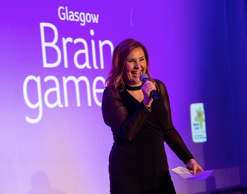 A woman holding a microphone, behind her is the Marie Curie logo and the words "Glasgow Brain game"