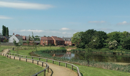 A dirt pathway with a residential area in the distance and some trees next to a lake on the right side of the pathway